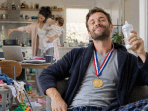 Homem com medalha no pescoço em primeiro plano, enquanto a mulher, ao fundo, segura o bebê, cozinha e fala ao telefone ao mesmo tempo