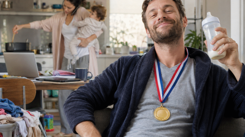Homem com medalha no pescoço em primeiro plano, enquanto a mulher, ao fundo, segura o bebê, cozinha e fala ao telefone ao mesmo tempo