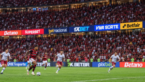 Jogadores disputam partida de futebol em estádio lotado, com diversas marcas de bets exibidas em placas publicitárias