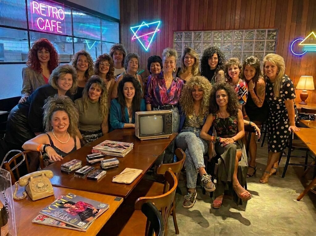 Imagem inspirada na trend dos anos 80, que recria fotos com estética dos anos 80. Um grupo de mulheres aparece com roupas e acessórios neon, ombreiras marcadas, polainas e penteados volumosos típicos da década