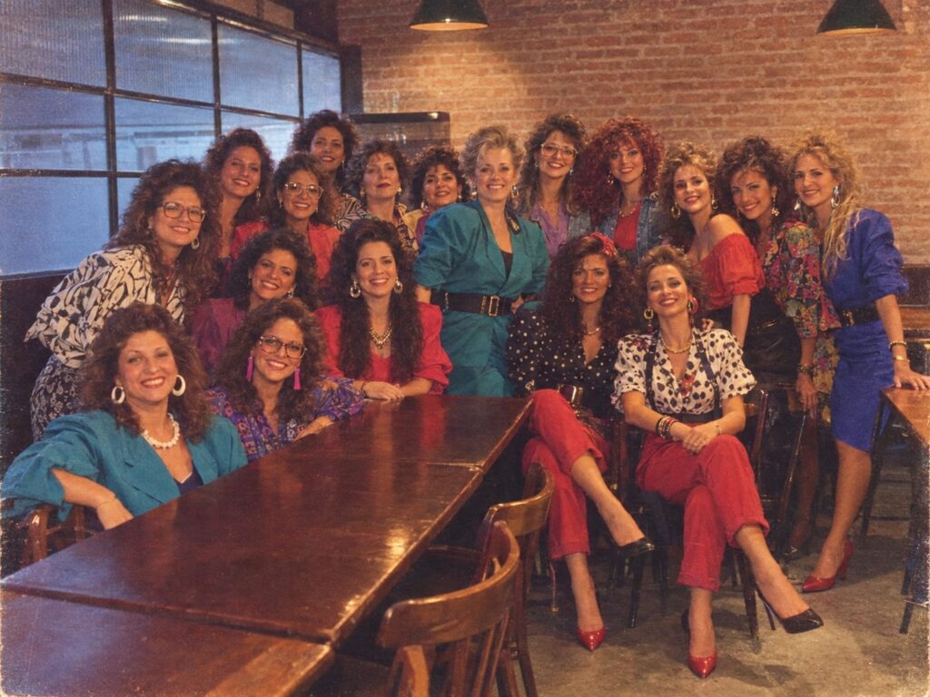 Imagem inspirada na trend dos anos 80, que recria fotos com estética dos anos 80. Um grupo de mulheres aparece com roupas e acessórios neon, ombreiras marcadas, polainas e penteados volumosos típicos da década