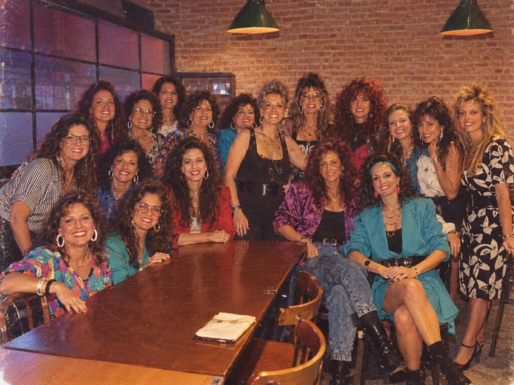 Imagem inspirada na trend dos anos 80, que recria fotos com estética dos anos 80. Um grupo de mulheres aparece com roupas e acessórios neon, ombreiras marcadas, polainas e penteados volumosos típicos da década