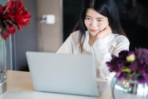 Mulher de camisa branca diante de um notebook, sentada à mesa com vasos de flores