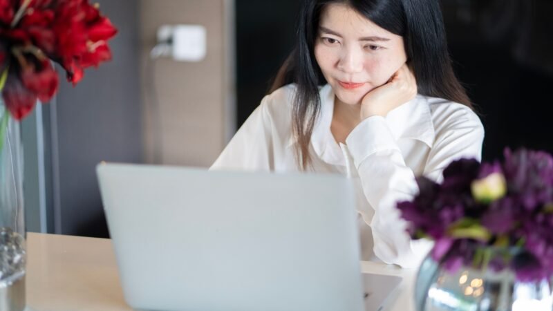 Mulher de camisa branca diante de um notebook, sentada à mesa com vasos de flores