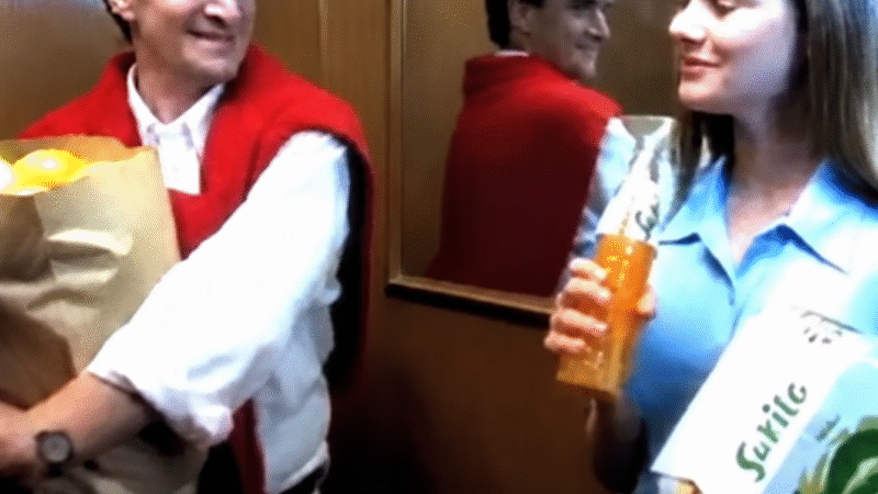 Roberto Arduim segura uma sacola de laranjas ao lado de Michelly Machri, que bebe Sukita, dentro de um elevador.